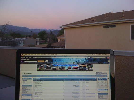 Laptop screen shows forum with vehicle banner; mountains and house in background