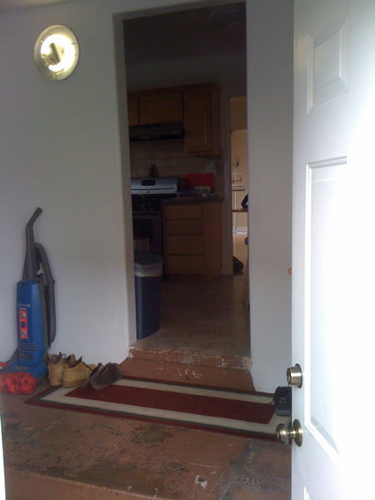 Interior view of a doorway leading to a kitchen, with a vacuum cleaner and shoes by the entrance