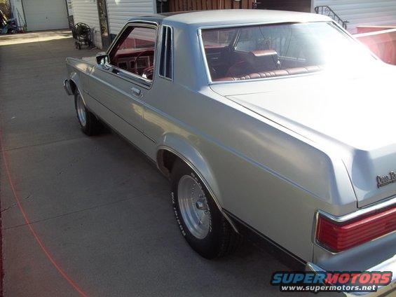 Silver coupe parked on a concrete driveway, rear quarter view. The car has a black vinyl roof and red interior