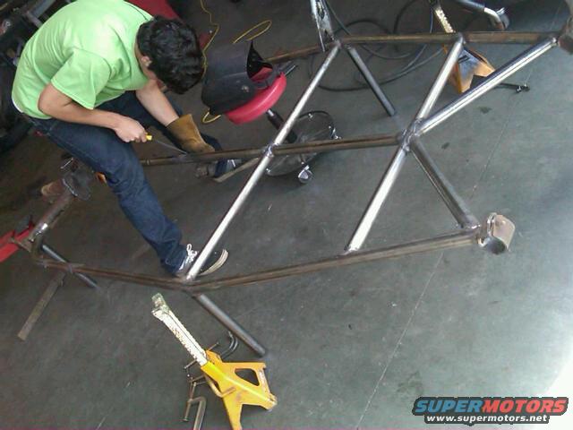 Person welding a custom metal frame in a garage, with a yellow jack stand visible