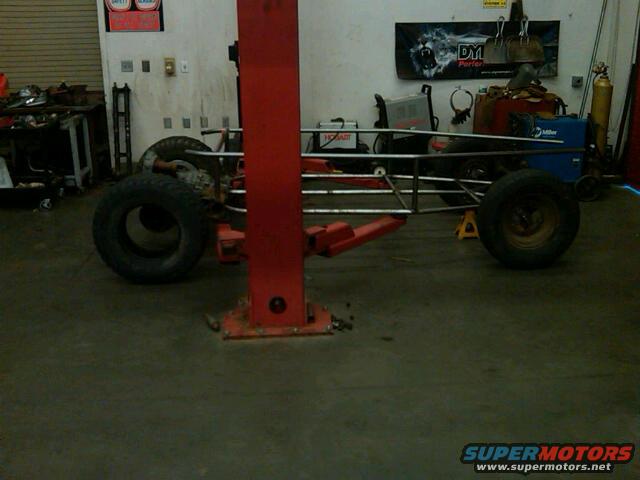 Bare metal frame of a custom car, possibly a dragster or kit car, on a lift in a garage