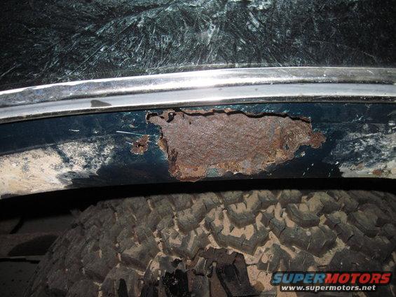 Dark green vehicle's rear wheel well showing significant rust damage and a chrome trim piece above