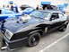 1956 Ford Fairlane - A heavily modified black coupe with a prominent supercharger is displayed at an outdoor event.