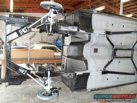 Underside of a 1956 Ford Fairlane chassis with suspension components visible, with another car in the background