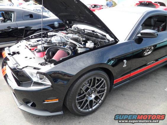 Black Ford Mustang GT500 with hood open showing engine and red air filter. Another Mustang is visible behind it