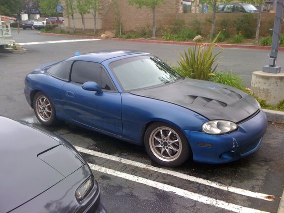 Blue Mazda Miata with a black vented hood parked next to a dark gray car