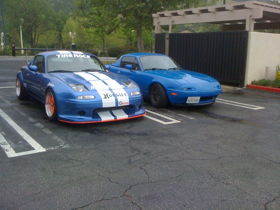 Two blue 1991 Mazda Miatas parked side-by-side, one with racing stripes and a spoiler