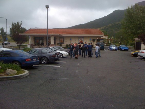 Group of people standing in a parking lot next to several parked cars, including a blue Mazda Miata and a dark gray Mazda Miata