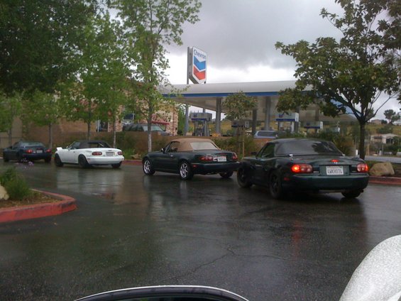 Three 1991 Mazda Miatas in a row at a wet gas station, rear view