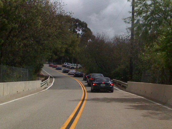 Cars driving on a winding road. The lead car is a dark convertible