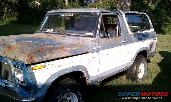 Rusty two-tone blue and white Ford Bronco with a dented front bumper and a missing headlight