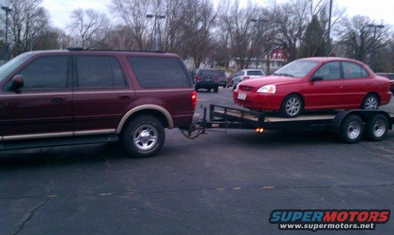 Maroon SUV towing a red sedan on a trailer. SUV appears to be a 2000 Ford Expedition