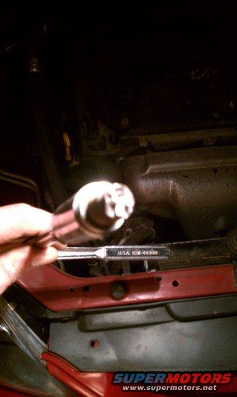 Hand holding a silver oxygen sensor with a black tip, over a red car part and dark engine components. A wrench with "USA ID# 44398" is visible