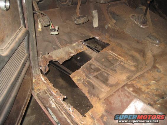 Interior view of a vehicle's floor pan with significant rust and holes, showing pedals and part of the door panel