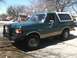 1989 Ford Bronco - This green Ford Bronco is parked on a street with houses and trees in the background.