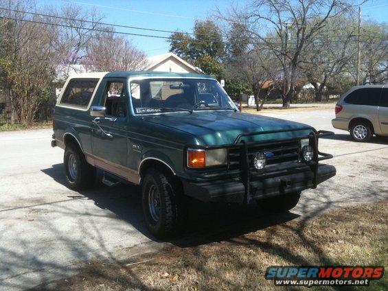 Green 1989 Ford Bronco parked on a street, with a tan minivan visible in the background