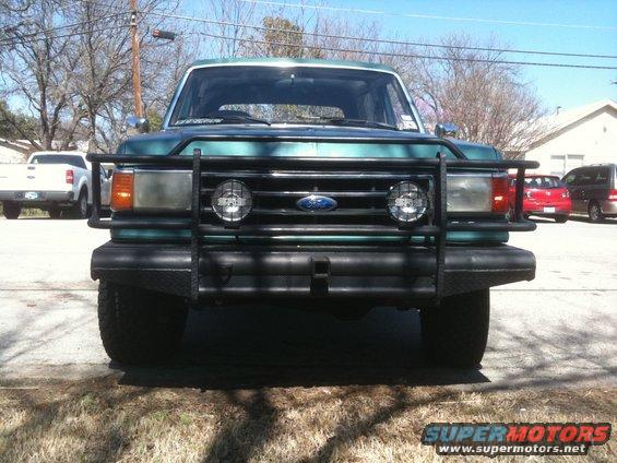 Green Ford Bronco with brush guard and aftermarket lights. White pickup truck in background