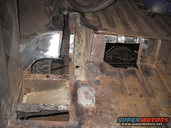 Underside of vehicle showing rusted floor pan and metal repairs. Tire visible in wheel well