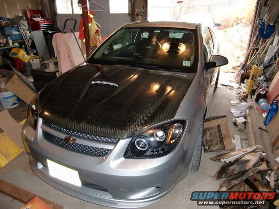 Silver sedan, possibly a Chevrolet Cobalt, with a carbon fiber hood and aftermarket headlights, parked in a cluttered garage