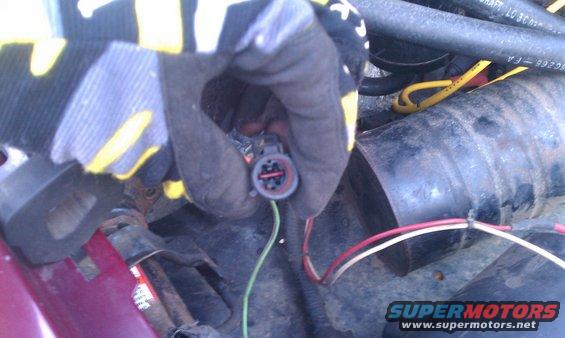 Gloved hand holding a gray electrical connector with red and green wires attached, near engine components