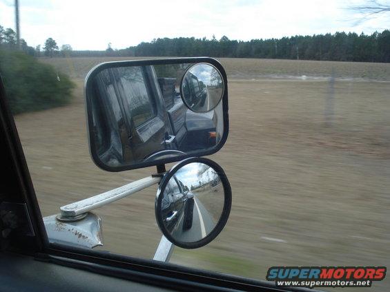Truck side mirror reflecting a road and trees, with a second smaller mirror below showing the road ahead
