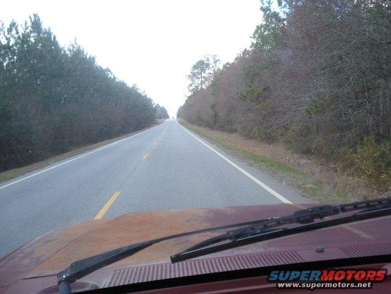 View from inside a vehicle looking over a rusty hood and windshield at a two-lane road lined with trees