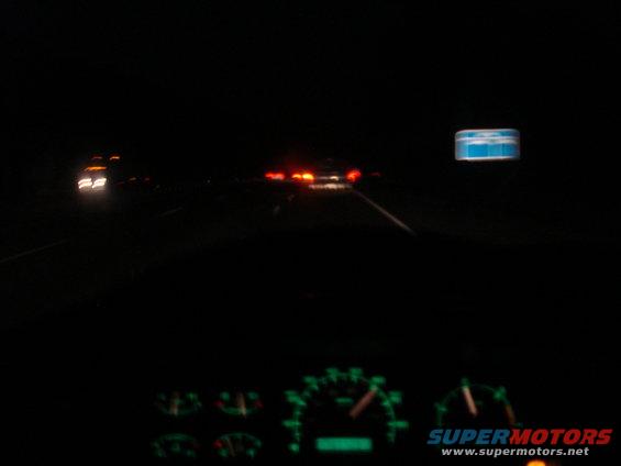 Dashboard gauges glow green at night, with blurry headlights and taillights of other vehicles visible on a highway