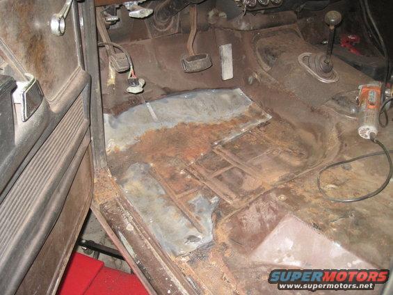 Interior floor of a vehicle with significant rust and patch panels, showing pedals and a shifter