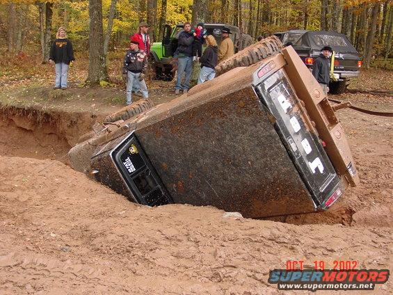 Mud-covered truck overturned in a dirt pit, with people watching