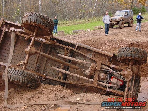 Overturned pickup truck chassis stuck in mud; another SUV is parked nearby