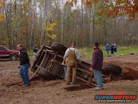 Off-road vehicle flipped on its side in mud with people watching. Red SUV in background