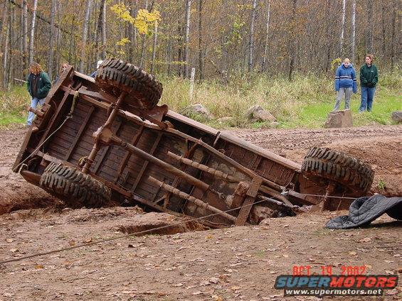 Overturned rusty truck stuck in dirt with large tires, people watching in background