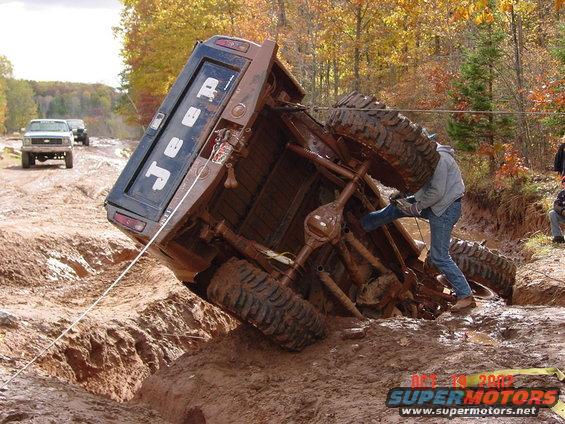 Rolled-over Jeep truck in mud with people attempting recovery. Another truck visible in background