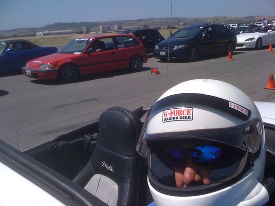 Driver wearing a white helmet with G-Force Racing Gear stickers, sitting in a 1991 Mazda Miata convertible. Other cars are visible on a track