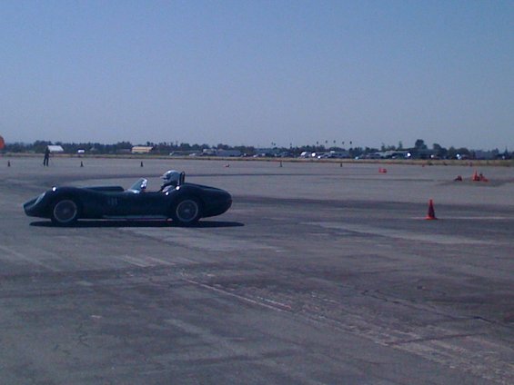 dark colored vintage race car with driver wearing helmet on a paved track with orange cones