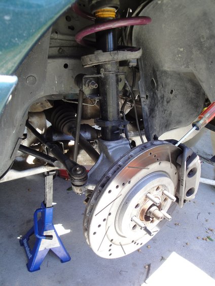 Green car's front suspension with a jack stand, perforated brake rotor, and caliper. The car appears to be from the early 2000s
