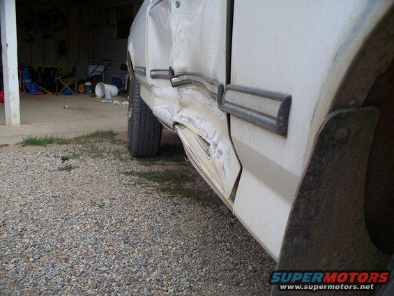 Side of a white truck with heavy damage to the door and lower body panel. Mud flap visible