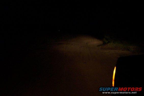 Headlight beam illuminates a dirt road at night, with a sliver of a vehicle visible on the right