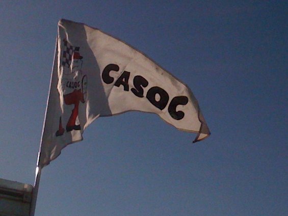 White flag with black text CASOC and a checkered flag logo against a blue sky