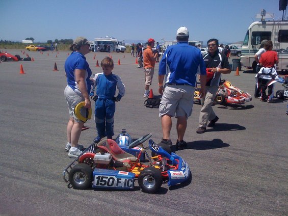 Go-kart with number 150 FIC on side, people in racing suits and casual clothes, RVs and other go-karts in background at track