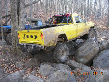 Yellow Jeep J-10 pickup truck with a roll cage, covered in mud, navigating large rocks