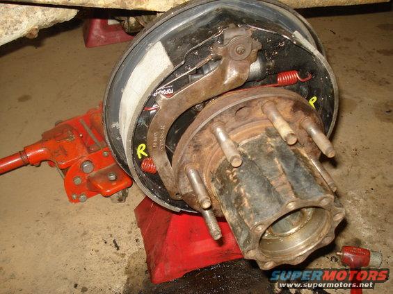 Close-up of a rusty brake drum and hub assembly with brake shoes and springs. A red jack is visible