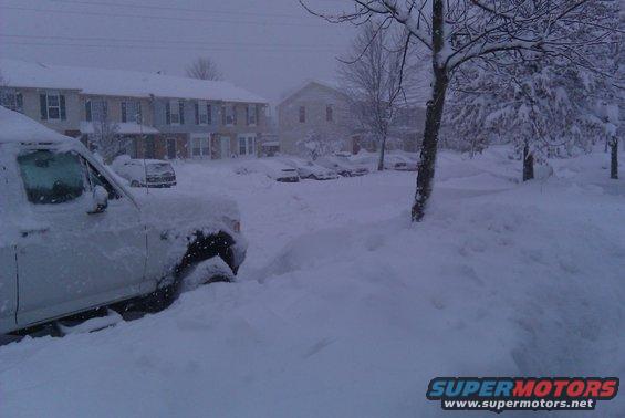 White SUV stuck in deep snow, front quarter view, with houses and other snow-covered vehicles in background