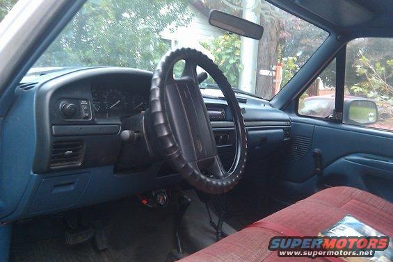Interior view of a truck cab showing the dashboard, steering wheel, and red bench seat. The dashboard has a blue plastic finish