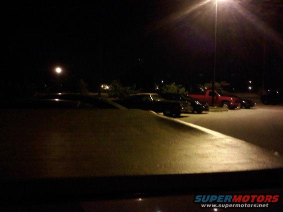 Several cars parked in a lot at night under a streetlamp, with a red truck visible