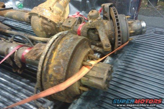 Two rusty truck axles with suspension components and brake rotors are strapped down in a truck bed