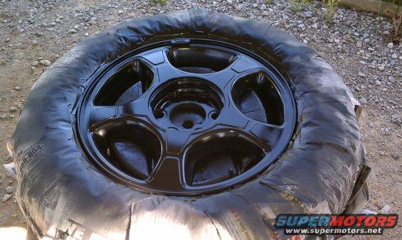 Black painted car wheel covered in plastic wrap, resting on gravel