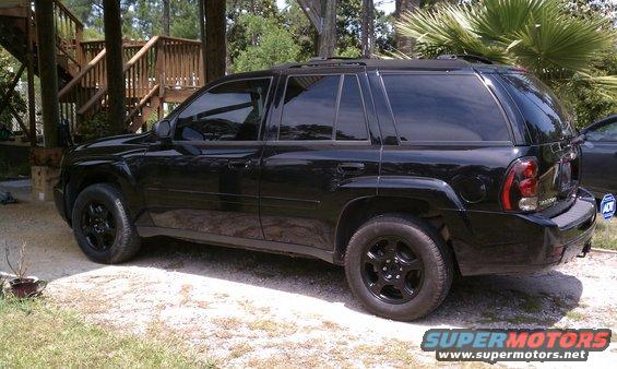 Black SUV parked on gravel, side view. Tinted windows, black wheels, roof rack