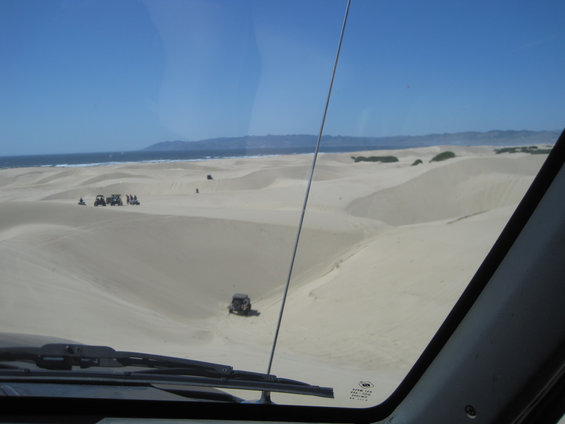 View from inside a vehicle looking out at sand dunes and the ocean, with several ATVs and a Jeep visible