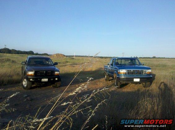 Blue 1992 Ford F-150 parked on a dirt road, front 3/4 view. Dark colored truck to the left